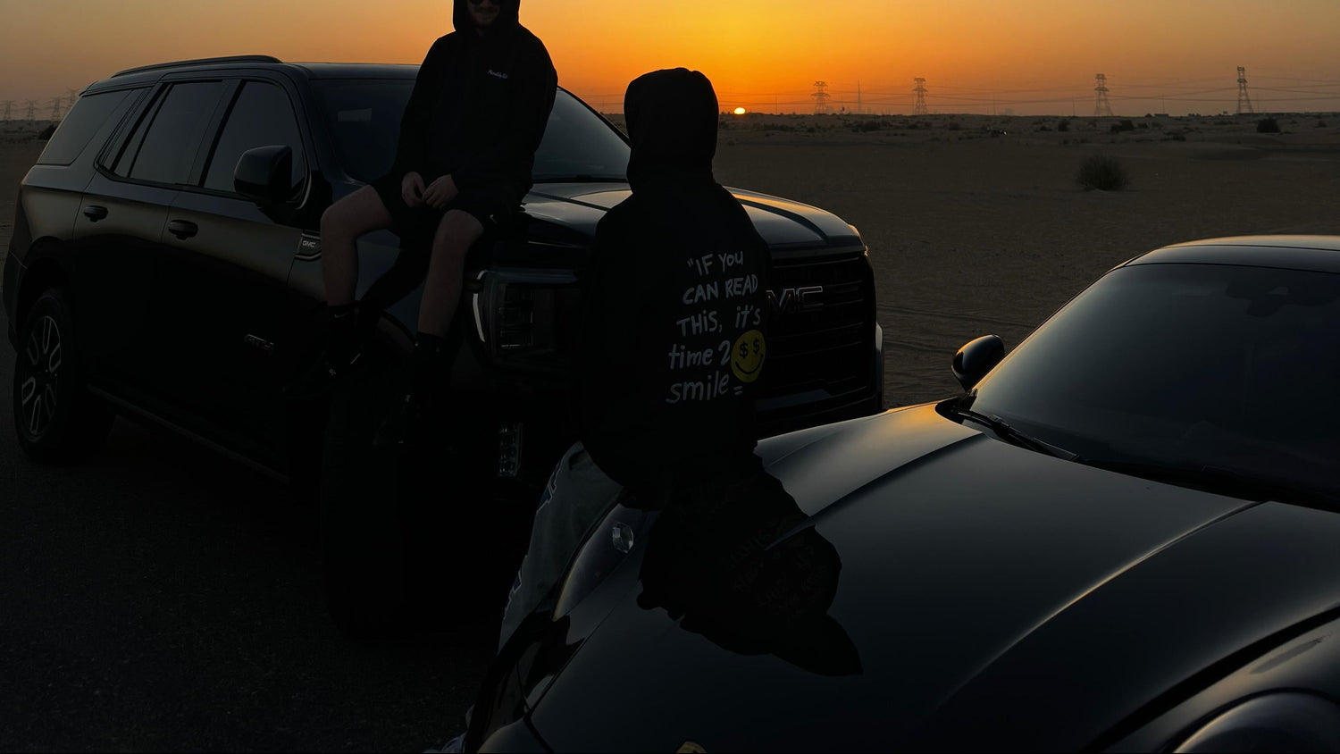 Two people sitting on a car in a desert at sunset